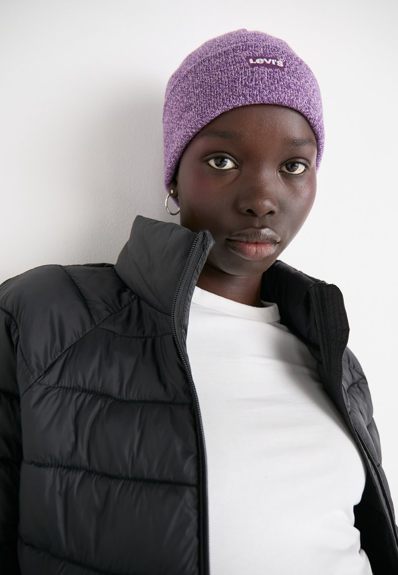 Gorro de punto morado con un logo blanco, combinado con una chaqueta acolchada negra y una camiseta blanca. Primer plano del gorro y la parte superior del cuerpo.