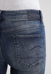 Jeans i denim med en mörkblå tvätt, prydda med en läderlogotyp på midjebandet och detaljer i sömmarna på bakfickan. Snittet är figursytt.