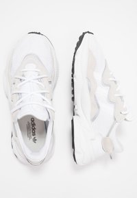 Zapatillas deportivas blancas y beige claro con texturas de malla y ante, que presentan una suela negra gruesa y líneas de diseño intrincadas.