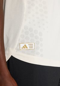 Bela mrežasta športna majica z pikčasto vzorcem, z zlatim logotipom in tremi črtami, prilegnjenega kroja, v kombinaciji s črnimi hlačami.