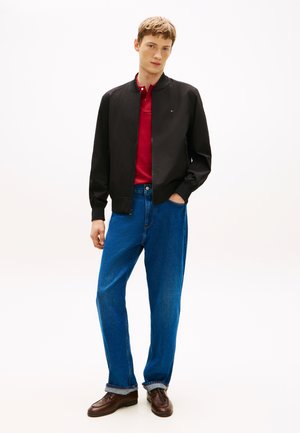 Schwarze Bomberjacke, rotes Poloshirt, blaue Jeans mit weitem Bein, braune Schuhe; glatter Stoff mit gerippten Bündchen, minimalistisches Design.