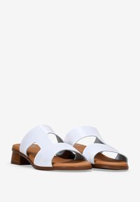Sandalias abiertas de piel blanca con detalles laterales recortados y un bajo tacón de madera marrón. Textura suave con un diseño minimalista.
