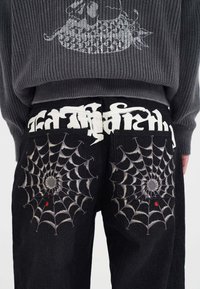 Black pants with embroidered gray spider webs and small red spiders on back pockets, worn with a gray sweater featuring a white fish design.