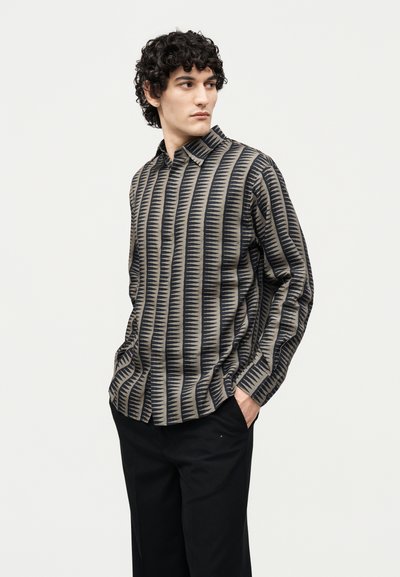 Άνδρας με κατσαρά μαλλιά που φοράει πουκάμισο με μακριά μανίκια και κάθετες μαύρες και μπεζ γεωμετρικές ρίγες, με τα χέρια στις τσέπες ενός μαύρου παντελονιού, κοιτάζοντας πλάγια.