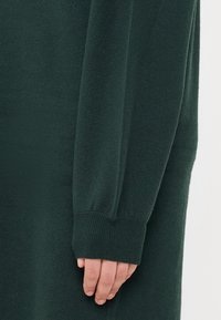 Mørkegrønn strikkegenser arm med ribbede mansjetter og myk tekstur, med en hånd som hviler mot stoffet.
