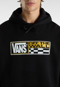 Zwarte hoodie met een groot Vans-logo in wit en geel, voorzien van een geruit motief. Zachte textuur en verstelbare trekkoordkap.