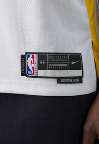 Bílý basketbalový dres s jemnou texturou, s černou nášivkou s logem NBA, velikost 44, a textem "ENGINEERED". Žluté akcenty na rameni.