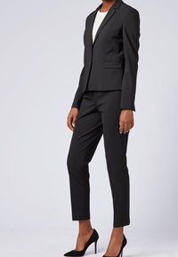 Black tailored suit featuring a slim fit blazer and matching trousers. Smooth fabric with subtle sheen, paired with black high-heeled shoes.