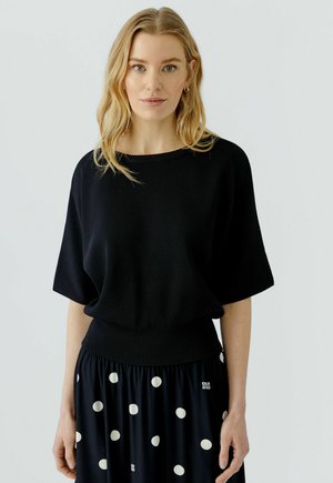 Vrouw met lang blond haar draagt een loszittende zwarte top en een zwarte rok met grote witte stippen, staand tegen een effen achtergrond.