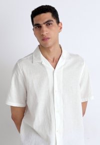 Chemise en lin blanche à manches courtes avec un devant à boutons, un col décontracté et une texture douce, portée par un mannequin se tenant devant un fond uni.