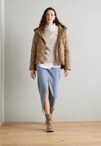 Veste matelassée marron sur un pull côtelé beige et une chemise blanche à col, associée à une jupe en denim clair avec une fente sur le côté et des bottines beiges.