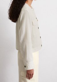 Lys beige linned cropped jakke med knaplukning, to brystlommer og en afslappet pasform. Knapdetaljer ved manchetterne tilføjer et pift.