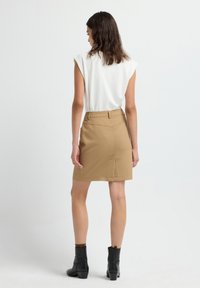 Vrouw in een mouwloze witte top, tan rok met een achteropening, en zwarte enkelboots, staande met de rug naar de camera tegen een witte achtergrond.