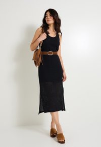 Vestido de ganchillo negro sin mangas con un dobladillo de longitud media y una abertura lateral, combinado con un cinturón marrón y un bolso con flecos.