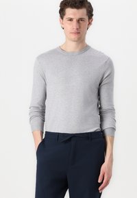 Hellgrauer Pullover mit langen Ärmeln aus glattem Stoff, mit einem gerippten Rundhalsausschnitt und Bündchen, kombiniert mit dunkelblauen Hosen.