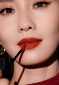 Gros plan sur une femme appliquant du rouge à lèvres mat rouge profond à l'aide d'un pinceau, avec un teint lisse et un maquillage léger pour les yeux. Des boucles d'oreilles en or sont présentes.