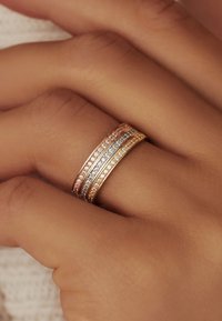 Ros�égoldring mit drei gestapelten Bändern; zwei Bänder sind mit kleinen klaren Steinen besetzt, und ein Band ist glatt. Strukturiert Hand im Hintergrund.