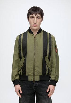 Mann mit kurzem dunklem Haar, der eine olivgrüne Bomberjacke mit schwarzen vertikalen Streifen und schwarze Jeans trägt, steht vor einem einfarbigen Hintergrund.