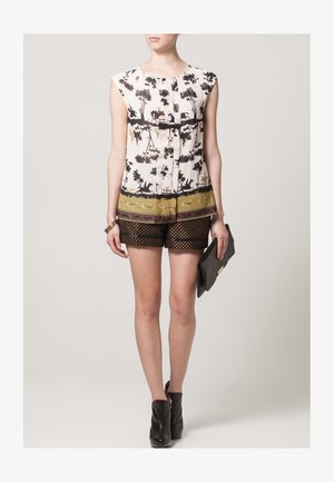 Vrouw draagt een mouwloze blouse met een zwart-beige natuur- en dierenprint, zwarte doorschijnende korte broek met polkadots, zwarte enkellaarzen en houdt een zwarte clutch vast.