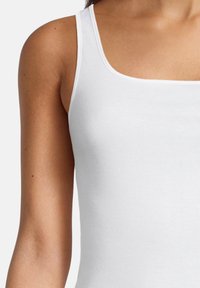 Woman wearing a white sleeveless tank top, showing her left shoulder and upper chest against a plain background.