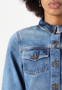Light blue denim shirt with a pointed collar, golden button closures, and two chest pockets with flap and button detail.