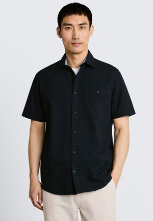 Mężczyzna ubrany w czarną teksturowaną koszulę na krótki rękaw na białą koszulkę, w połączeniu z jasnymi spodniami, stojący na jednolitym tle.