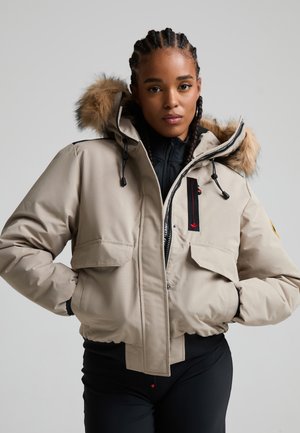 Femme aux cheveux tressés portant une veste d'hiver beige avec capuche doublée de fourrure et un pantalon noir, debout les mains dans les poches sur un fond uni.