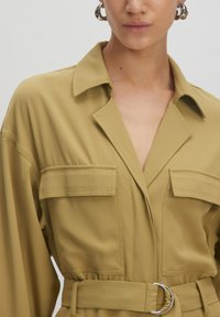 Olivgrünes Hemdkleid mit Gürtel, Kragen, kurzen Ärmeln, zwei Brusttaschen und einem silbernen runden Schnallendetail in der Taille.