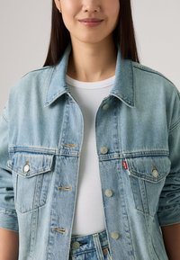Veste en denim bleu clair avec deux poches poitrine, boutons-pression en argent et col classique. Portée sur un t-shirt blanc à côtes.