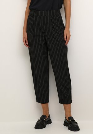 Frau trägt schwarze, knöchellange Nadelstreifenhose kombiniert mit schwarzen Loafers, steht vor hellem, einfarbigem Hintergrund.