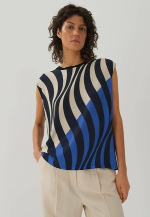 Top à manches courtes en bleu, marine et crème avec des motifs ondulés. Tissu doux avec une coupe décontractée et un col rond, porté avec un pantalon clair.
