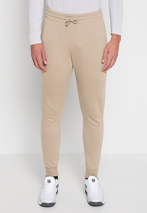 Pantalon de jogging beige slim à taille coulissante, porté avec des chaussures de sport blanches et une chemise blanche à manches longues.