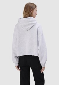 Kobieta o jasnobrązowych włosach, ubrana w jasnoszarą skróconą bluzę z kapturem i czarne jeansy, stoi tyłem na jednolitym jasnym tle.
