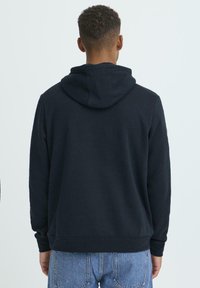 Tmavomodrá mikina s kapucňou z mäkkej látky, vybavená sťahovacou šnúrkou na kapucni, dlhými rukávmi a rebrovanými manžetami a spodným lemom. Pohľad zozadu bez loga.