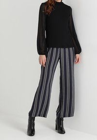 Svart långärmad topp i genomskinligt material med hög krage, kombinerad med navy och vita randiga vida byxor. Svarta högklackade stövlar fulländar looken.