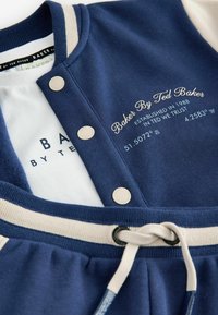 Tumesinine ja kreemjas värvitoonis vabaaja outfit bomber-stiilis jackeetiga, millel on nööbid ja broderitud brändi detailid, valge särgi peale kihtidena.