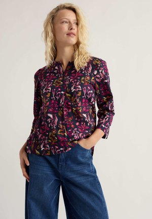 Szőke nő sötétkék, virágos, hosszú ujjú blúzban és bő kék farmerral, egyik kezét zsebében tartva, balra nézve áll.