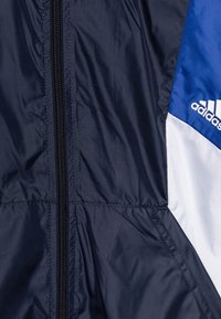 Marineblauwe windbreaker met contrasterende witte en blauwe panelen, voorzien van een ritssluiting aan de voorkant, getextureerde stof en een Adidas-logo op de borst.