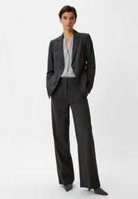 Grau maßgeschneiderter Anzug mit einem taillierten Blazer mit einem einzelnen Knopf, weiter Hosen und einer hellgrauen Bluse darunter. Glatte Stoffoberfläche.