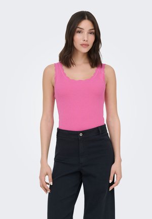Femme portant un débardeur côtelé rose vif sans manches avec des bords festonnés et un pantalon noir à taille haute, debout devant un fond clair.