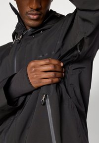 Zwarte waterdichte jas met een hoge kraag, ritszakken en een mesh voering. Het model past een binnenzak aan met een aansluitende manchet.