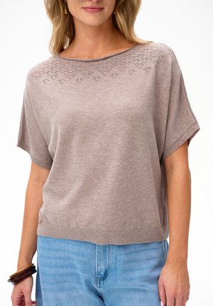 Kobieta w jasnobrązowym, krótko rękawowym, dzianym topie z koronkowym dekoltem i jasnoniebieskich dżinsach, uzupełniona brązowymi bransoletkami.