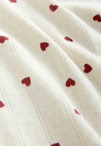 Kermavalkoinen kangas, jossa on teksturoitu kuvio punaisista sydämistä ja ohut pystysuora raitakuosi. Pehmeä rakenne, kevyt ja hengittävä.