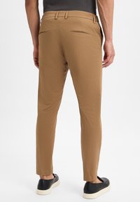 Man in een slim-fit bruine broek en zwarte instappers, staand met ontspannen handen langs zijn zij tegen een witte achtergrond.