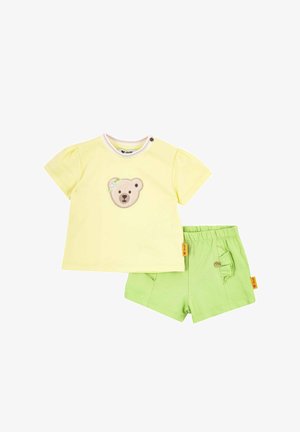 Gelbes kurzärmeliges Kleinkind-Shirt mit gesticktem Bärengesicht und grüne Shorts mit elastischem Bund und aufgesetzten Taschen mit Rüschen und Knöpfen.