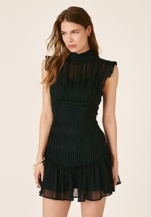 Mujer con largo cabello castaño, vistiendo un minivestido negro plisado sin mangas con dobladillo con volantes, mirando ligeramente hacia un lado sobre un fondo liso.