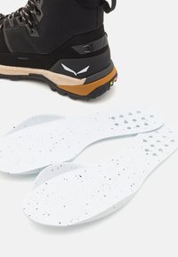 Bota de senderismo con parte superior negra y suela color marrón, acompañada de plantillas blancas con manchas negras y recortes para una mayor transpirabilidad.