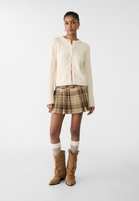 Beige strikket cardigan med knaplukning, parret med en ternet plisseret nederdel i brun og beige, samt brune ruskind cowboy støvler.
