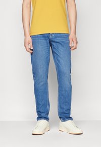 Ljust blå jeans med en rak bendesign, denimmaterial och subtila blekningar. Bärs med en gul T-shirt och vita sneakers.