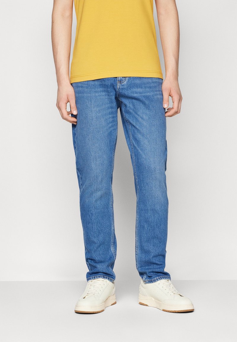 Ljust blå jeans med en rak bendesign, denimmaterial och subtila blekningar. Bärs med en gul T-shirt och vita sneakers.